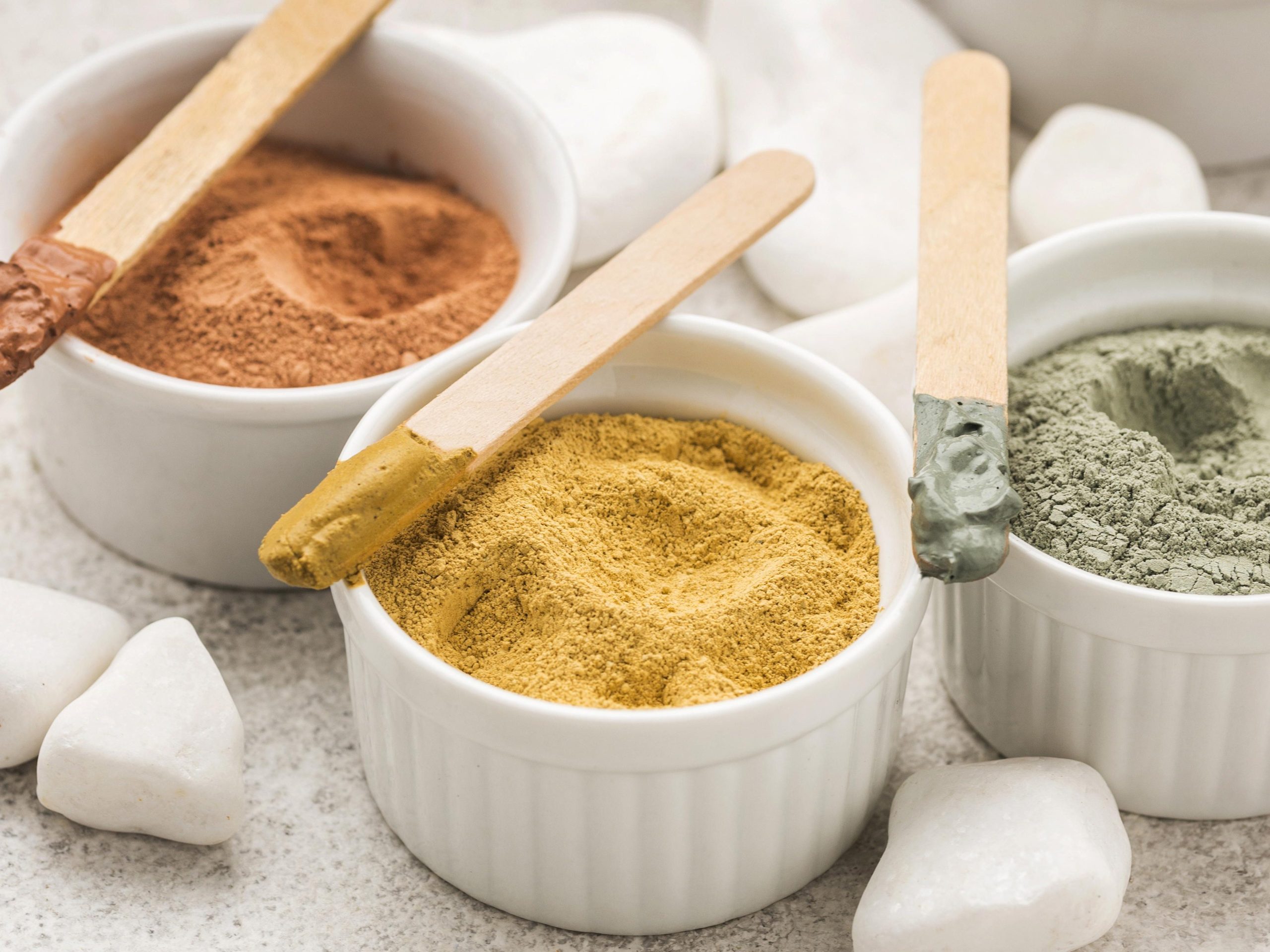 Види косметичної глини для обличчя у білому посуді з дерев'яними палочками