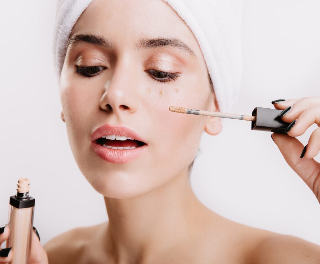 Beautiful girl after shower posing on white background. Woman puts concealer to hide bruises under eyes.