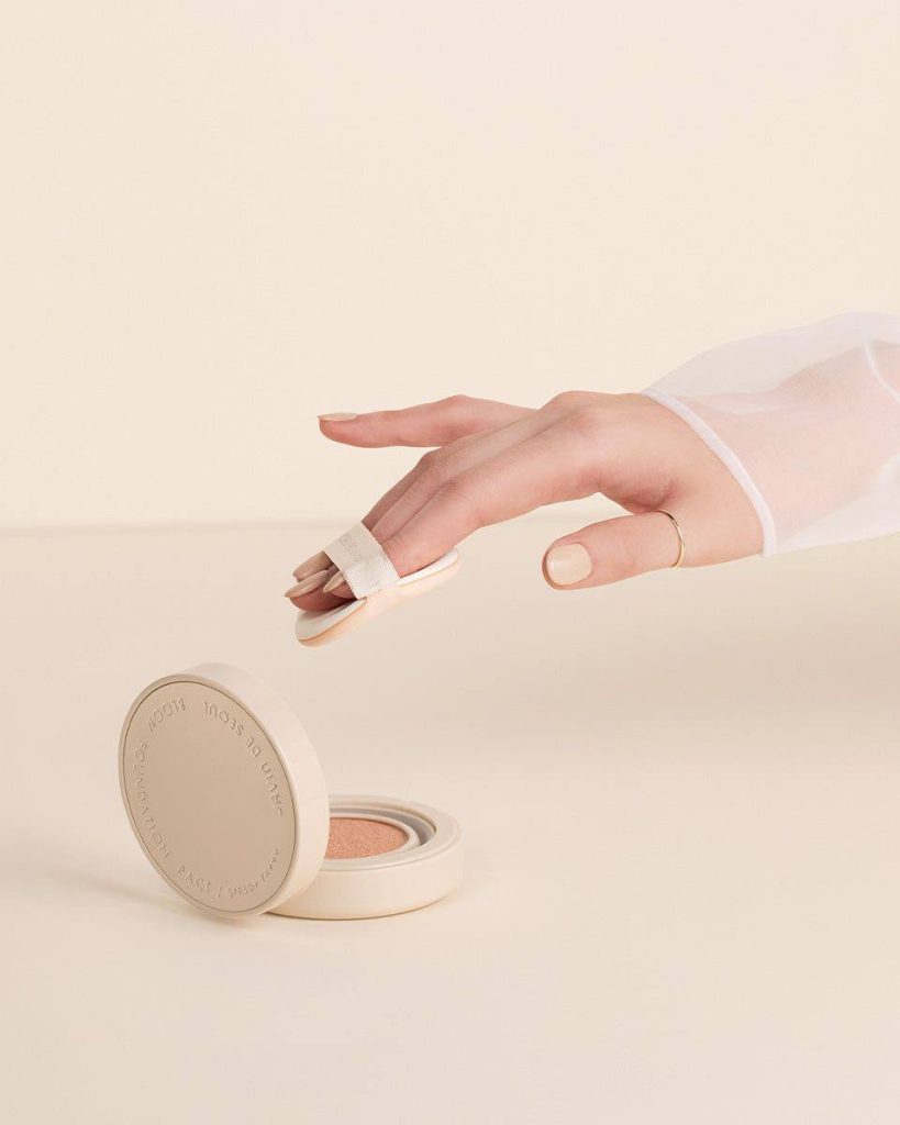 ДІвчина у білій прозорій блузці набирає спонжиком кушон з упакування