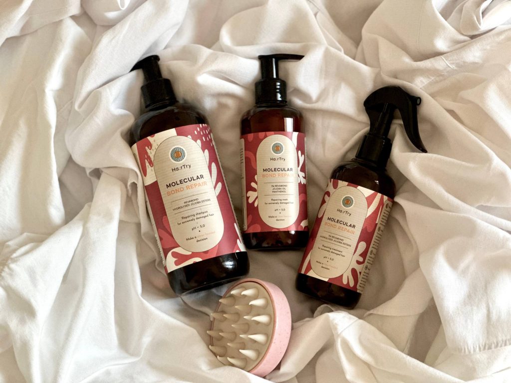 Средства из новой линейки HairTry — Molecular Bond Repair на белой простыне, рядом с ними — силиконовая щетка для массажа и мытья головы